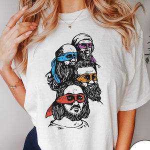 May include: White t-shirt with a graphic of four men wearing ninja turtle masks. The men have beards and are stacked on top of each other. The text "WHITE" is visible in the bottom right corner of the image.