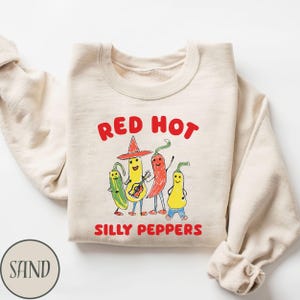 May include: A sand-colored sweatshirt with the words "RED HOT" above a cartoon illustration of four anthropomorphic peppers. The peppers are playing musical instruments and wearing hats. The words "SILLY PEPPERS" are below the illustration.
