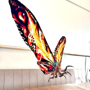 Puede incluir: Una escultura de insecto alado grande, colorida, con un cuerpo detallado y texturizado y patas largas y afiladas. La escultura está montada sobre una base de madera y está pintada en tonos de rojo, naranja, amarillo y negro.