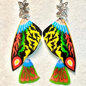 May include: A pair of handmade earrings featuring a butterfly wing design. The earrings have a silver-colored butterfly charm at the top, with colorful wings in shades of yellow, black, red, green, and blue. The bottom of the earrings have a green, red, and blue triangle shape.