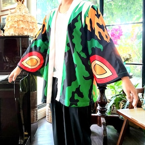 Peut inclure: Une veste de style kimono colorée avec un motif audacieux de formes vertes, orange et noires. La veste a de larges manches et une doublure blanche. Les manches présentent un motif rouge, jaune et orange. Le vêtement est porté sur un haut blanc et un pantalon noir.