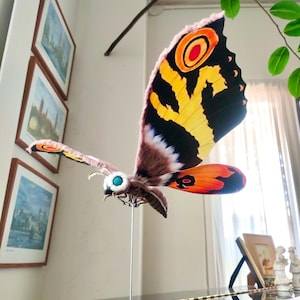 May include: A detailed, hand-crafted model of the Mothra moth from the Godzilla franchise. The model is made of fabric and features intricate details, including textured wings with black, yellow, orange, and red patterns, and large, blue eyes.