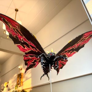 May include: A black and red monster with wings, resembling a moth, with yellow accents. The monster has a spiky head and red eyes. The monster is posed in flight with its wings spread wide.