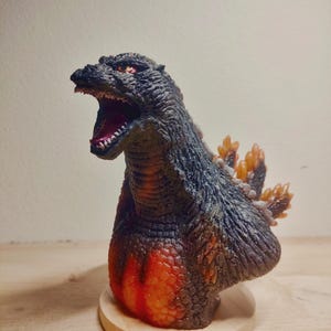 May include: A black and orange Godzilla bust figurine with a roaring expression. The figurine is made of plastic and has a detailed sculpt. The bust is mounted on a wooden base.
