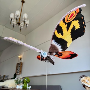 May include: A large, colorful moth model with black, yellow, orange, and red wings. The moth is mounted on a wooden stand and is in a flying position.
