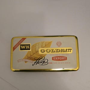 以下が含まれることがあります： 金色の縁取りが付いた長方形の銀色の缶。缶には「GOLD BLATT」と「EXPORT」の文字が金と赤で書かれており、タバコの葉のイラストも描かれています。「WK-Tabak Bremen-Berlin」の文字も確認できます。