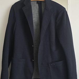 May include: A navy blue blazer featuring a grey interior lining, two front buttons, and two patch pockets. The lower left pocket displays the letters A|X in a subtle tonal embroidery. The garment is displayed on a wooden hanger.
