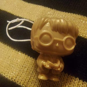 Puede incluir: Una figurita de plástico dorada de Harry Potter con gafas redondas y sosteniendo una varita mágica. La figurita es un adorno navideño con una cuerda blanca atada.