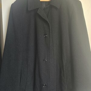 May include: A solid black coat featuring a classic collar, long sleeves, and a front button closure with three visible black buttons. The garment includes two side pockets and is displayed hanging on a wooden hanger, showcasing a simple design.
