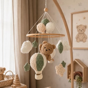Puede incluir: Un móvil colgante para guardería con un aro de madera central y elementos de ganchillo suspendidos que incluyen un oso de peluche marrón, un globo aerostático en verde y crema, nubes blancas y estrellas verdes y crema, unidos con hilos blancos.