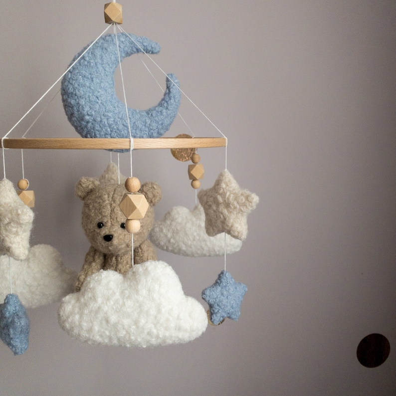 May include: A baby mobile with a wooden ring, blue and white crocheted stars, clouds, and a teddy bear. The mobile also features wooden beads and a crescent moon.