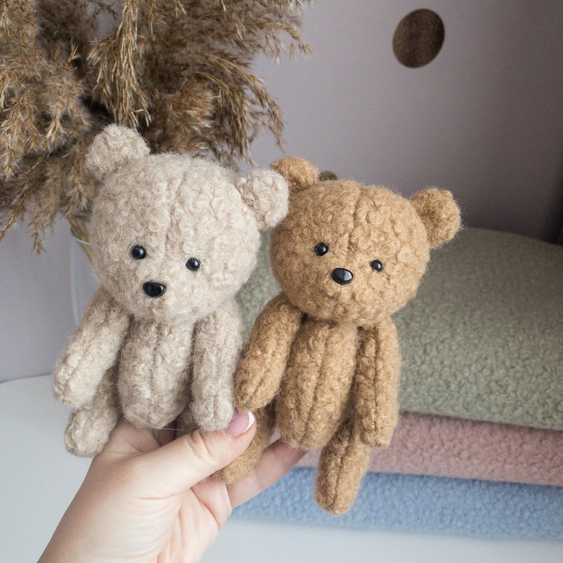 May include: Two plush teddy bears, one beige and one brown, with black eyes and noses. The bears are sitting on a stack of fabric.