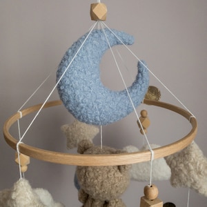 May include: A wooden baby mobile with a blue crescent moon, two beige clouds, a beige star, and a beige teddy bear. The mobile is suspended from a wooden ring.