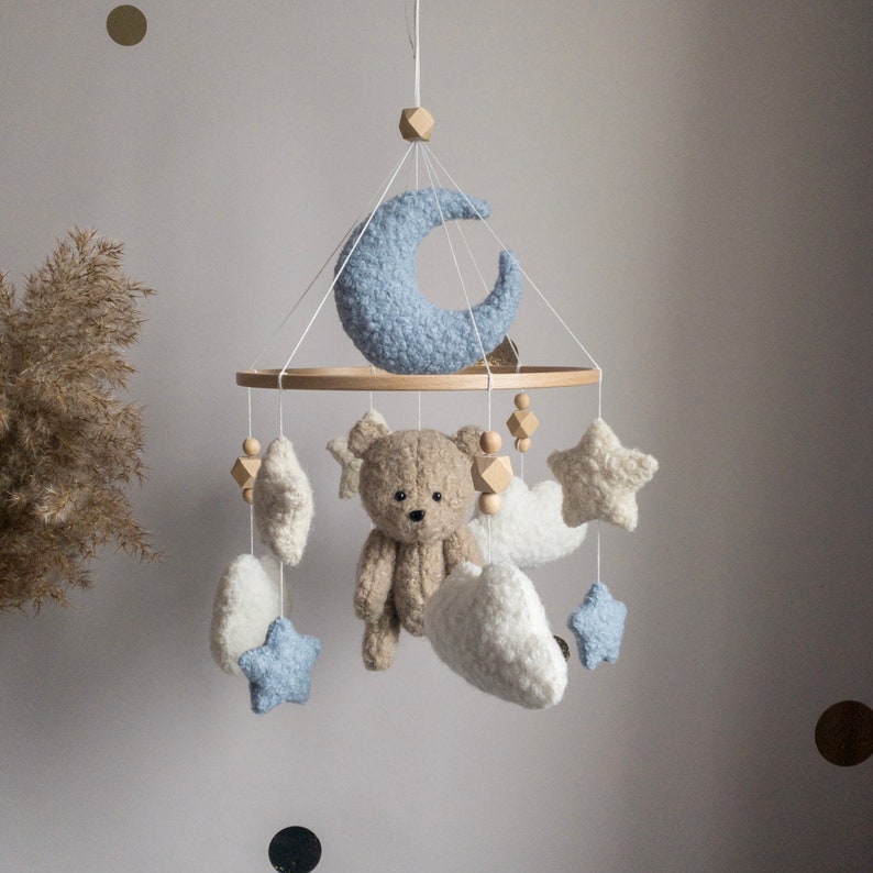 May include: A baby mobile with a wooden ring, a blue crescent moon, a brown teddy bear, white clouds, and blue stars.