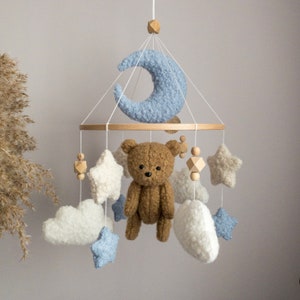 May include: A baby mobile with a brown teddy bear, white clouds, blue stars, and a blue crescent moon. The mobile is made of wood and fabric.