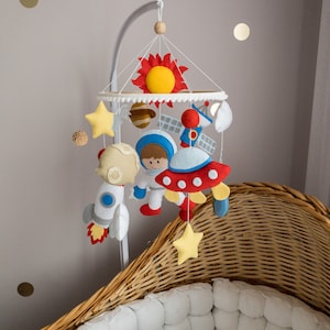 Op de afbeelding: Een witte en rode vilten astronaut-mobiel met een gele zon, sterren en planeten die aan een witte metalen ring hangen. De mobiel hangt boven een rieten wieg.