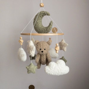 May include: A nursery mobile with a teddy bear, stars, clouds, and a crescent moon. The mobile is suspended from a wooden ring by white strings. The bear is tan, the moon and stars are green, and the clouds are white.