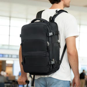 Puede incluir: Mochila de viaje negra con múltiples compartimentos, cremalleras y correas ajustables. La mochila tiene un asa superior y se lleva en la espalda. La persona lleva una camiseta blanca y vaqueros.