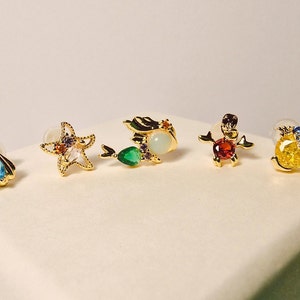 Puede incluir: Cinco pendientes de tono dorado con piedras preciosas de colores y diseños marinos. Los pendientes presentan una concha, una estrella de mar, una sirena, un cangrejo y un pez.