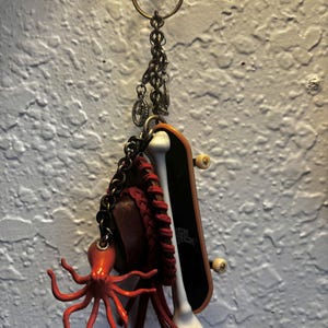 Könnte beinhalten: Ein Schlüsselanhänger mit einem schwarzen Miniatur-Skateboard, einem weißen knochenförmigen Anhänger, einer dunkelbraunen geflochtenen Lederschlaufe und einer leuchtend orangefarbenen Krakenfigur, befestigt an einem Metallring mit bronzefarbener Kette.