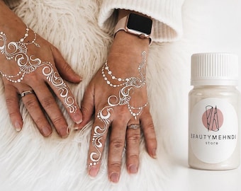 Può includere: Un piccolo flacone di plastica bianca con un'etichetta che riporta la scritta BEAUTYMEHNDI store, accompagnato da due mani decorate con intricati motivi all'henné bianco caratterizzati da linee sinuose, disegni floreali e catenelle di perline.