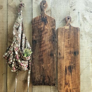 May include: Two tall, rectangular wooden cutting boards with dark, weathered grain and circular handle holes at the top, displayed alongside a brown fabric apron featuring a floral pattern and a small pocket containing a green artificial plant.