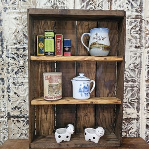 May include: A rustic, wooden shelf unit displays vintage kitchen items. The shelves hold spice tins, a blue and white floral mug, a small creamer, a baking powder tin, and two small, white cow planters with black spots. The unit is made of dark wood.