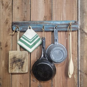May include: A rustic kitchen display featuring a wooden cutting board, a green and white pot holder, a black cast iron pan, a gray metal pot, and a wooden spoon. The items hang from a blue-painted wooden rack against a wooden wall.