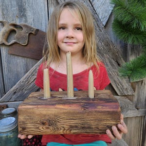 Puede incluir: Un portavelas rústico de madera con tres velas encendidas de color crema. El soporte está hecho de un trozo de madera rectangular y envejecido, que muestra la veta de la madera. Las velas son altas y delgadas, con goteo de cera.