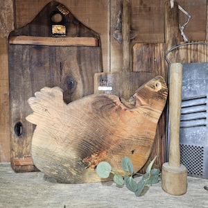 May include: A collection of rustic wooden cutting boards and kitchen tools. The centerpiece is a chicken-shaped cutting board, with other rectangular and uniquely shaped boards in the background. A wooden mallet and grater are also visible.