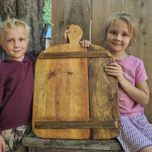 May include: A large, rectangular wooden cutting board with a handle. The board is made of several planks of wood, with darker wood accents. Two children are holding the board, smiling at the camera.