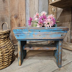 Peut inclure: Un petit tabouret rectangulaire en bois avec une finition en peinture bleue vieillie, doté d'un plateau plat, de quatre pieds robustes et d'une étagère de support inférieure. Un bouquet de fleurs artificielles roses et violettes est posé dessus.