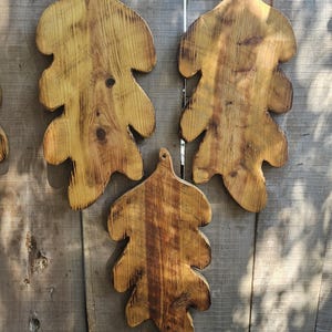 May include: Three wooden serving boards shaped like oak leaves, featuring a natural light brown wood grain with darker scorched edges and a small hole at the top of each board for hanging. The boards have a smooth, polished finish.