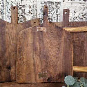 May include: Several wooden cutting boards with handles are displayed. The boards are made of dark brown wood with visible grain patterns. One board has a metal plate attached. The boards are arranged in a slightly overlapping manner.