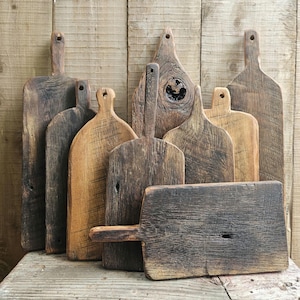 Könnte beinhalten: Eine Sammlung rustikaler Holzschneidebretter mit Griffen. Die Bretter variieren in Form und Größe und zeigen natürliche Holzmaserung und Farbtöne von Braun und Beige. Einige Bretter haben ein Loch im Griff zum Aufhängen. Die Bretter sind auf einer Holzoberfläche angeordnet.