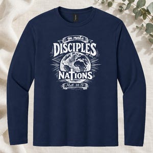 May include: A navy blue long-sleeve shirt featuring a white graphic print of the Earth with a cross in the center. The text reads: go make DISCIPLES OF ALL NATIONS Matt. 28:19. The design includes radiating lines behind the globe.