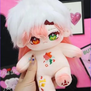 May include: A plush doll with white fluffy hair, pink and red accents, and large, detailed eyes. The doll has a small embroidered design on its chest and is held by a hand. The doll's hair is white and pink, and the eyes are green and red.