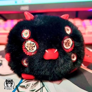 Pode incluir: Um bicho de pelúcia preto com chifres, nariz e pés vermelhos. O bicho de pelúcia tem vários detalhes circulares vermelhos com caracteres chineses. A criatura tem uma textura fofa e um design de desenho animado. O logotipo G4MBLITZ está no canto inferior esquerdo.