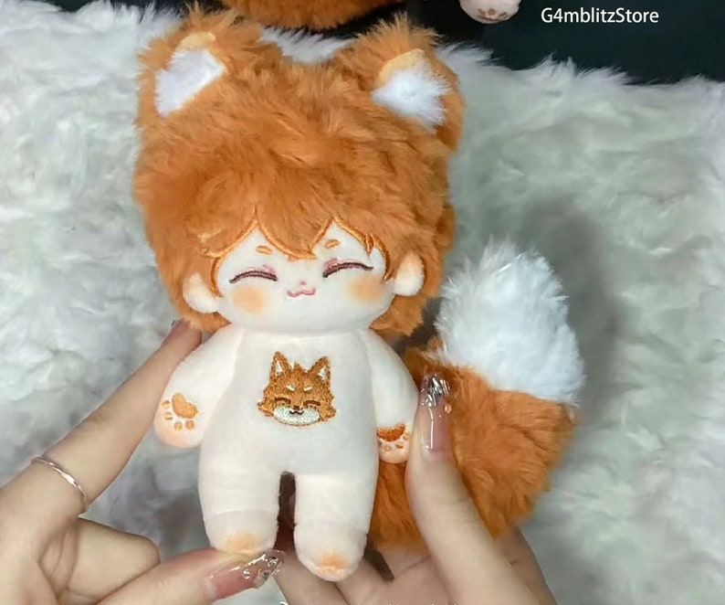 K&ouml;nnte beinhalten: Ein Pl&uuml;schtier-Fuchs mit orangefarbenem Fell, wei&szlig;en Ohren und einem wei&szlig;en Schwanz. Der Fuchs tr&auml;gt ein wei&szlig;es Outfit mit einem kleinen gestickten Fuchs auf der Vorderseite. Das Spielzeug hat ein s&uuml;&szlig;es, cartoonartiges Gesicht mit rosigen Wangen.