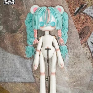 Può includere: Una bambola snodabile bianca con capelli in lana azzurra acconciati in due lunghe trecce con accenti rosa. La bambola presenta occhi a bottone blu, piccole orecchie da orso e un supporto in metallo. Il testo G4MBLitz è visibile nell'angolo.