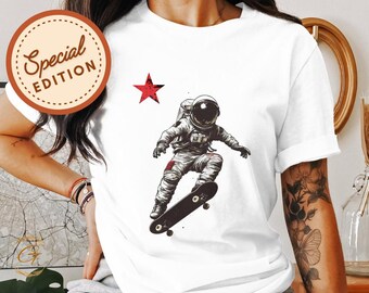 Puede incluir: Camiseta blanca con una estrella roja y una ilustración en blanco y negro de un astronauta haciendo skate. El astronauta lleva un casco y un traje espacial. La tabla de skate es negra con ruedas rojas.