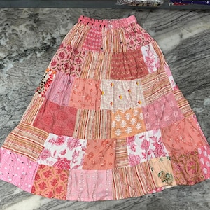 Pode incluir: Uma saia patchwork em tons de rosa, pêssego e branco. A saia apresenta uma variedade de padrões florais e geométricos, com cós franzido. A saia é feita de vários quadrados de tecido costurados.