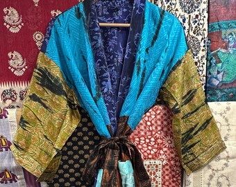 May include: A colorful patchwork robe with a tie closure, featuring shades of blue, green, and brown. The robe is hanging on a wooden hanger, with various patterned fabrics in the background.