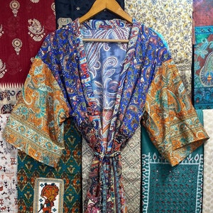 Puede incluir: Una bata de seda con estampado paisley azul, naranja y negro, con mangas largas y cinturón de lazo. La bata está colgada de una percha de madera frente a una pared de telas estampadas de colores.