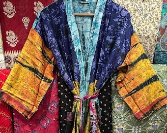 May include: A colorful silk kimono robe with a tie belt. The robe is made of patchwork fabric with different patterns and colors, including blue, yellow, orange, and red. The robe is hanging on a hanger.