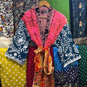 May include: An embroidered kimono in vibrant colours, hanging on a wooden hanger. The kimono features pink, blue, and orange panels with white stitching. The background displays various patterned fabrics.