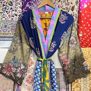 May include: A colorful, patterned kimono robe with long sleeves, hanging on a wooden hanger. The robe features a mix of floral, geometric, and abstract designs in shades of blue, gold, pink, and green. The background displays various textiles.