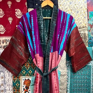 May include: A long, colorful robe with a tie belt. The robe is made of silk and has a tie-dye pattern in shades of pink, purple, blue, and green. The robe is hanging on a hanger in front of a wall with various patterned fabrics.
