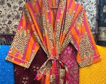 May include: An orange and pink striped robe with tiger print accents. The robe is hanging on a wooden hanger and tied with a matching belt. The robe is displayed against a backdrop of various patterned fabrics.