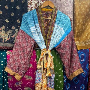 May include: A vibrant patchwork kimono robe in shades of blue, red, and yellow. The robe features a mix of patterned fabrics and long sleeves. It hangs on a wooden hanger, with a backdrop of various patterned textiles.